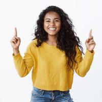 Cheerful optimsitic young female student with curly dark hairstyle, yellow sweater, smiling and laughing happily, pointing fingers up, showing friends link to site or copy space, white background.